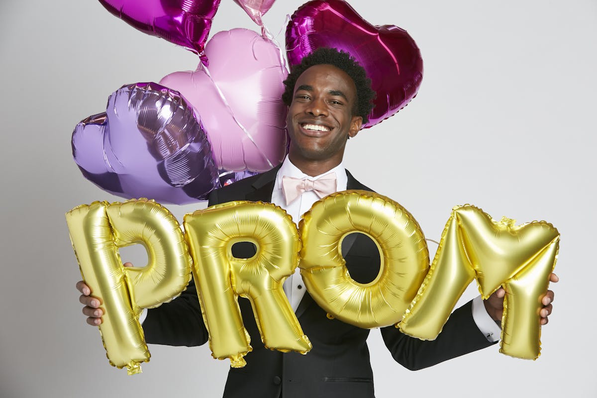 High school guy in black prom tuxedo with blush bow tie showing how to prompose with balloons.