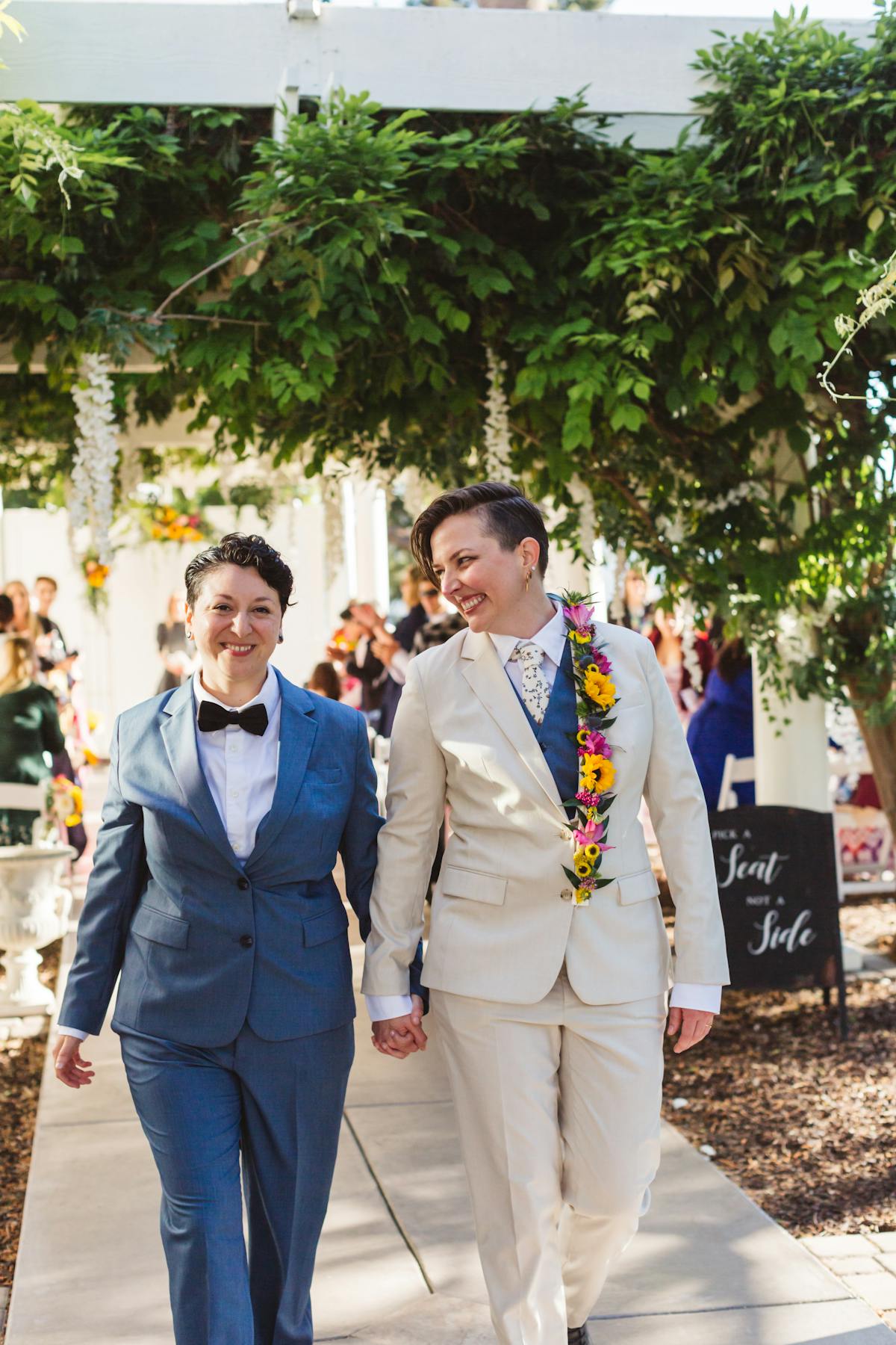 Newlywed brides in suits holding hands walking down the aisle wearing light blue and tan alternative bride outfits.