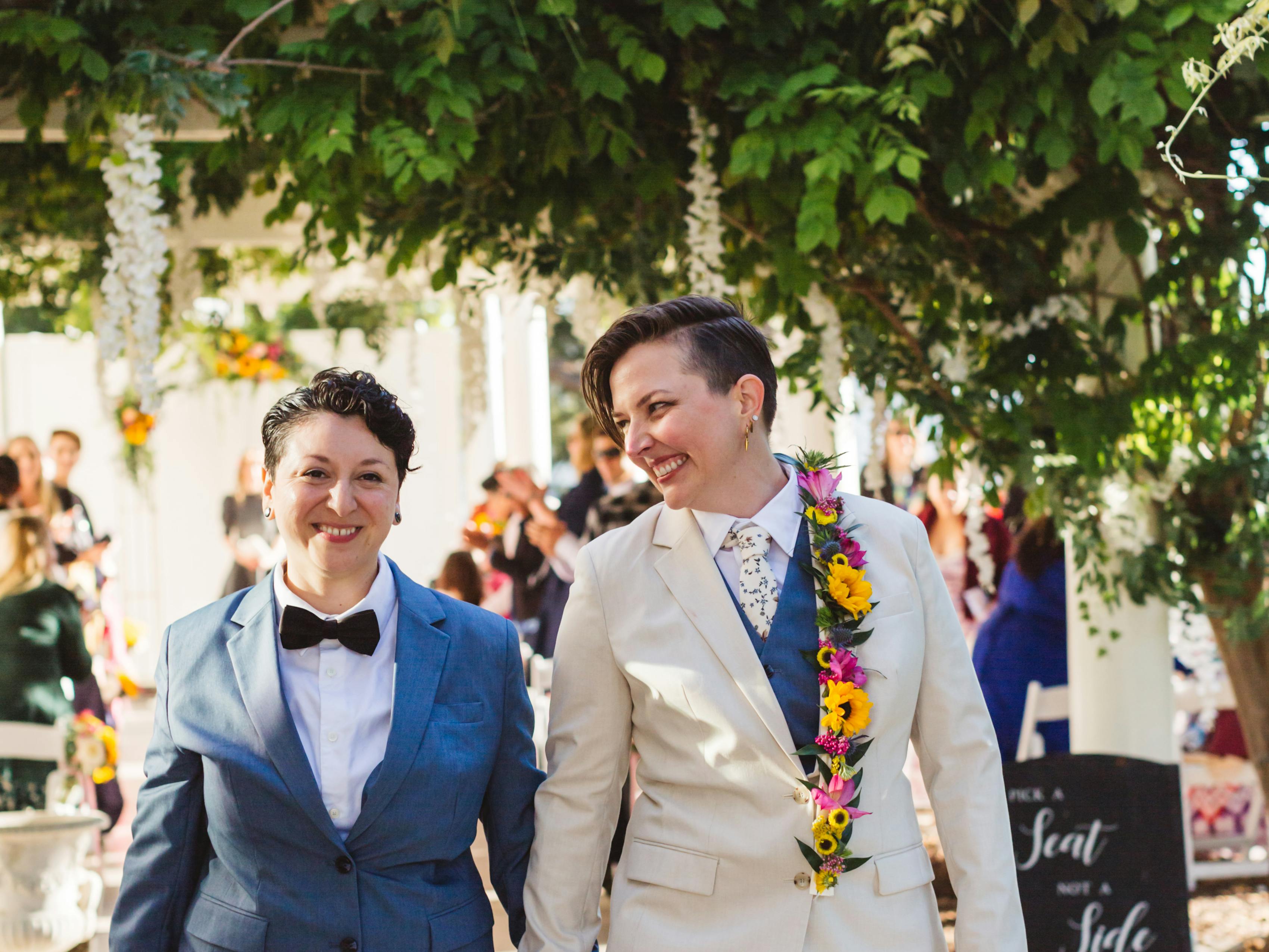 Newlywed brides in suits holding hands walking down the aisle wearing light blue and tan alternative bride outfits.