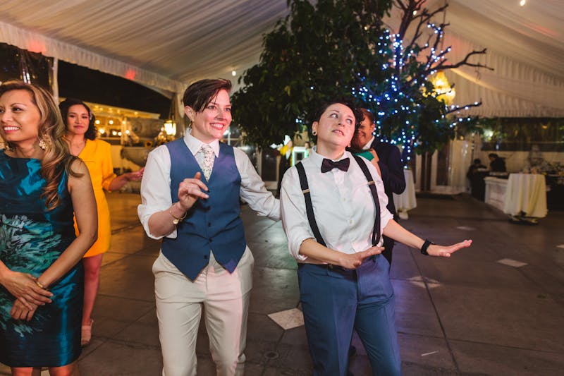 2 androgynous brides dancing in women's wedding suits, 1 with a women's suit vest and 1 wearing suspenders.