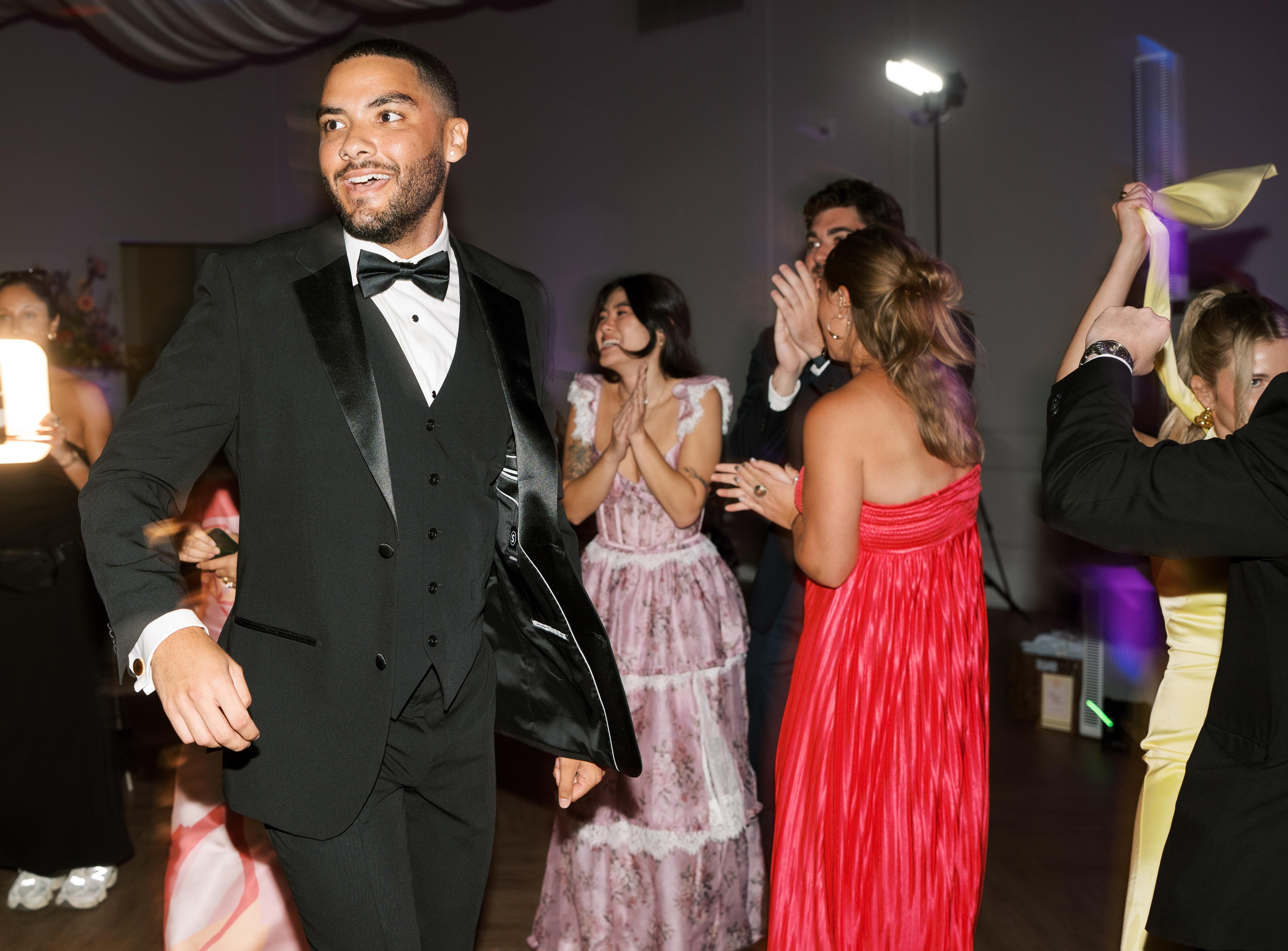 Man wearing a black three-piece tuxedo with satin lapels and a bow tie dancing at a formal event while guests in colorful dresses celebrate behind him.