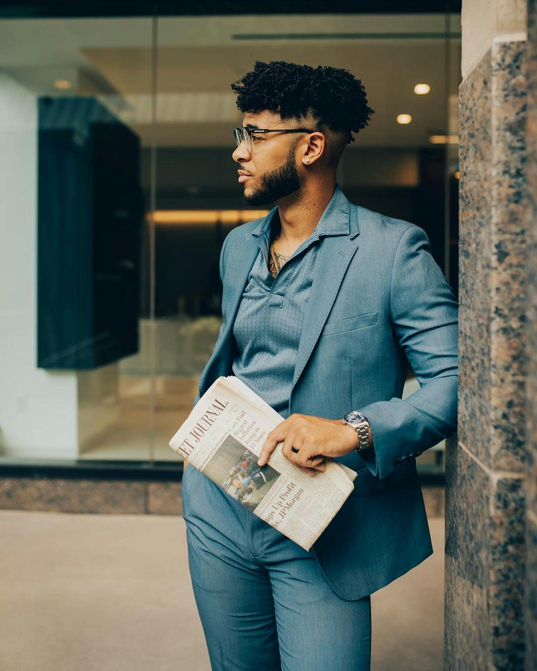 Cool menswear outfit for work with a light blue men's blazer, polo shirt, blue suit pants, and glasses in industrial shopping center.