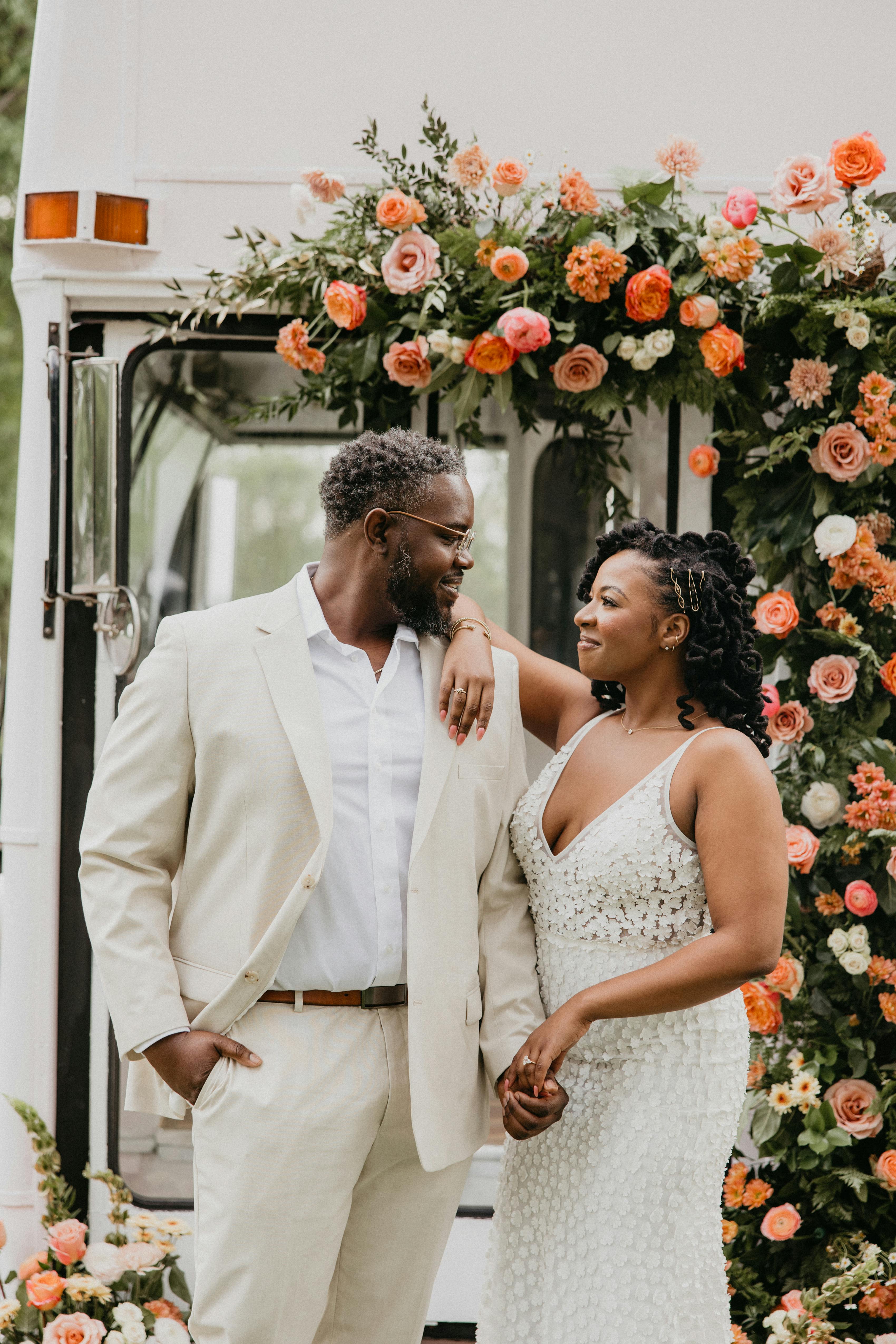 Groom wearing casual styled tan suit for men holding hands with bride wearing v-neck wedding dress in front of floral wedding arch
