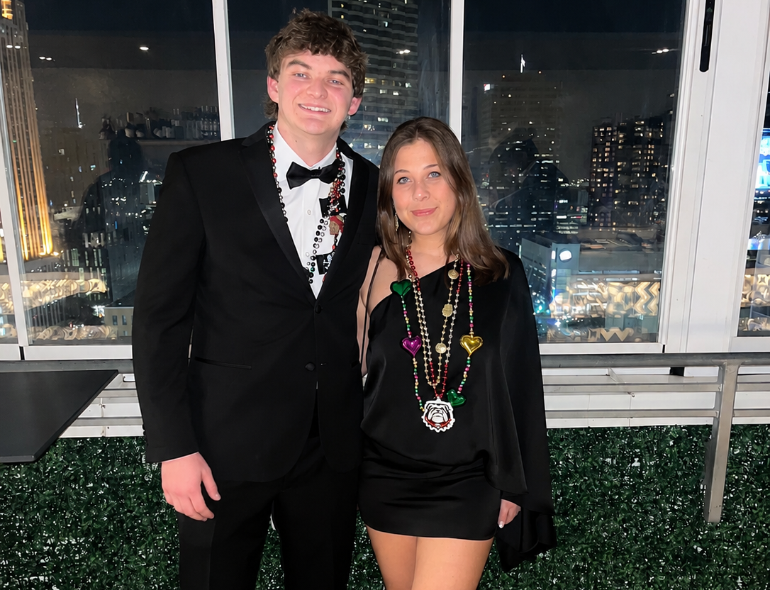 Couple posing at a fraternity formal with a nighttime city skyline behind them, the man in a black tuxedo and bow tie and the woman in a black mini dress, both wearing colorful Mardi Gras beads.