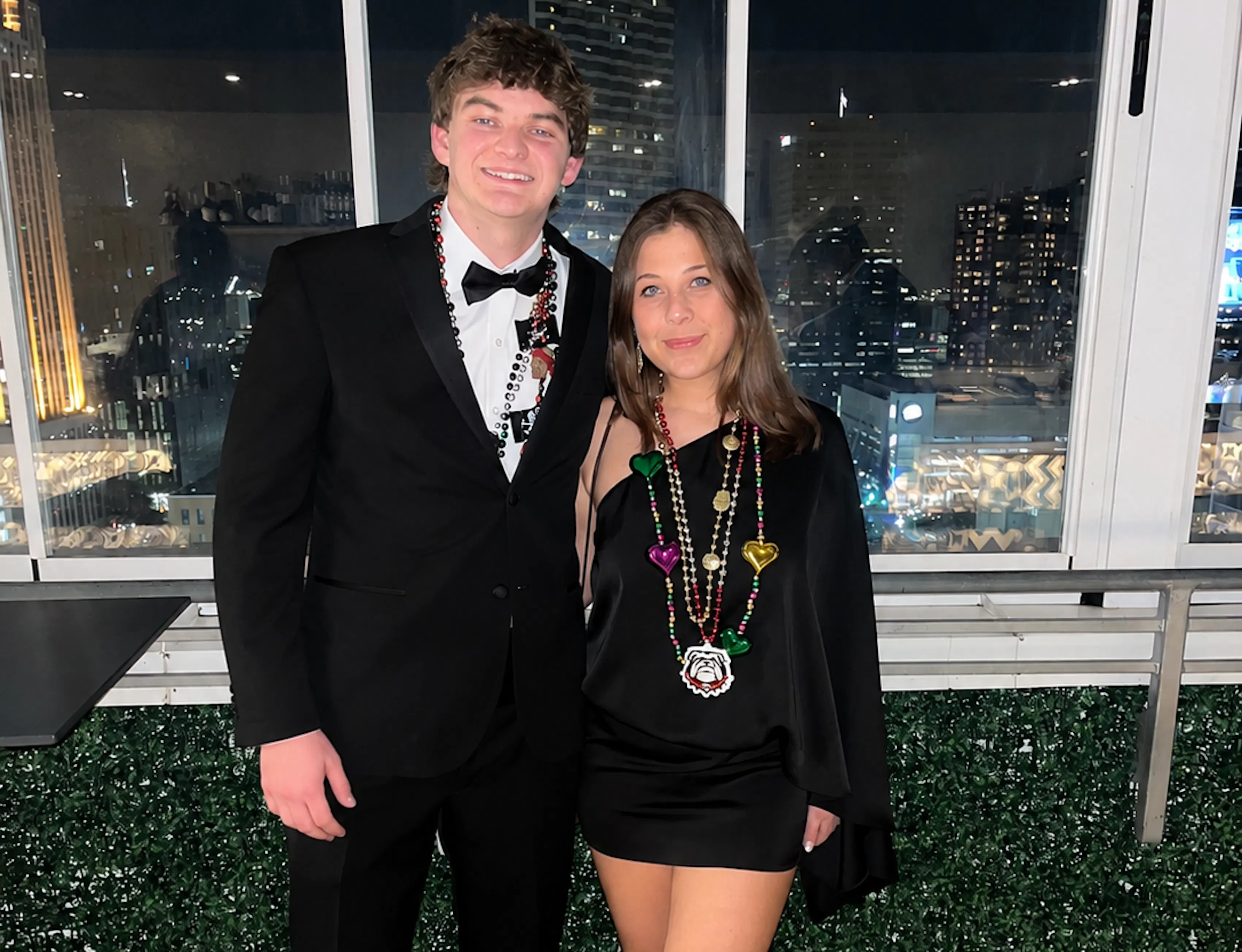Couple posing at a fraternity formal with a nighttime city skyline behind them, the man in a black tuxedo and bow tie and the woman in a black mini dress, both wearing colorful Mardi Gras beads.