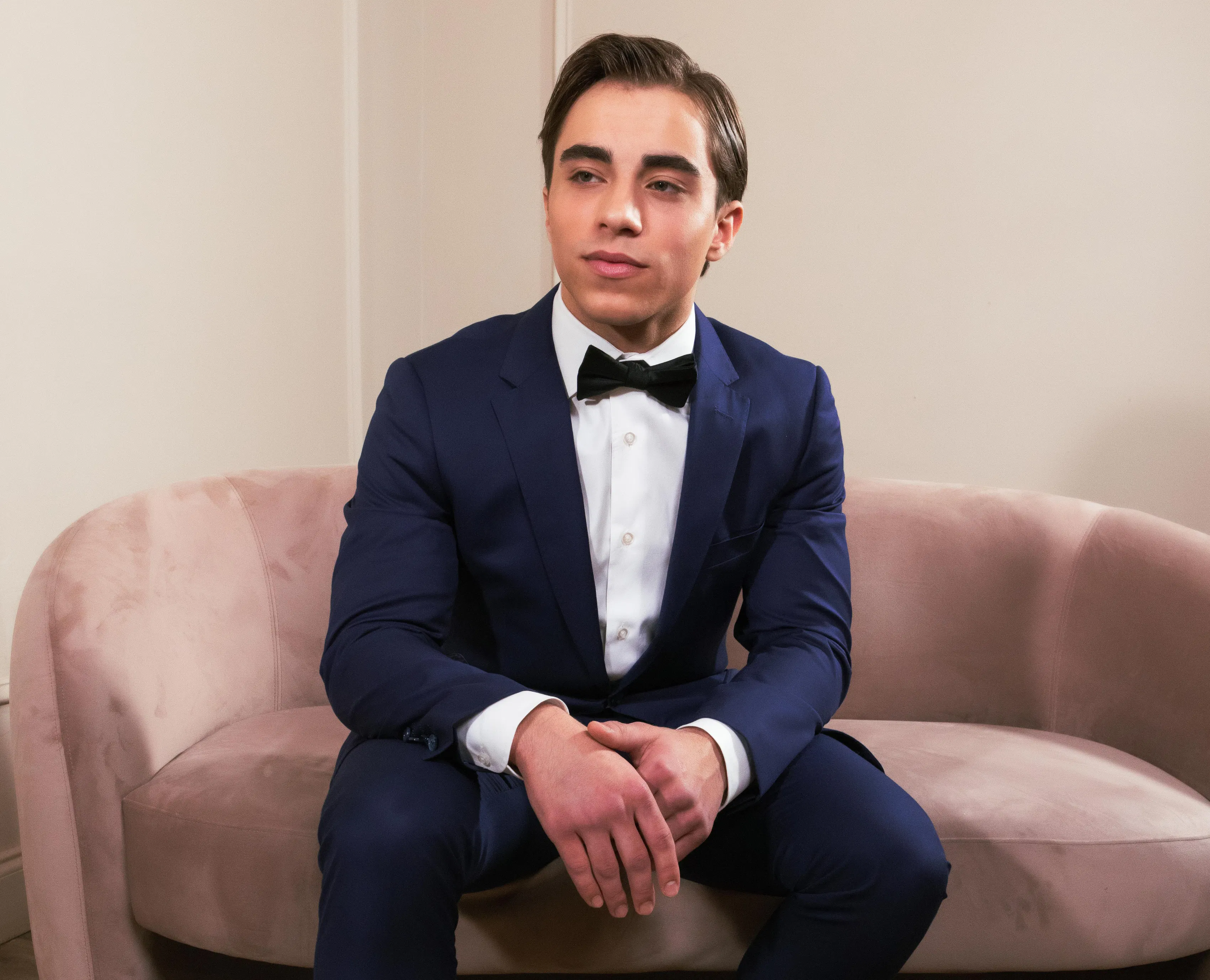 Short man wearing a fitted navy blue suit, white dress shirt, and black bow tie while sitting on a blush pink velvet sofa.