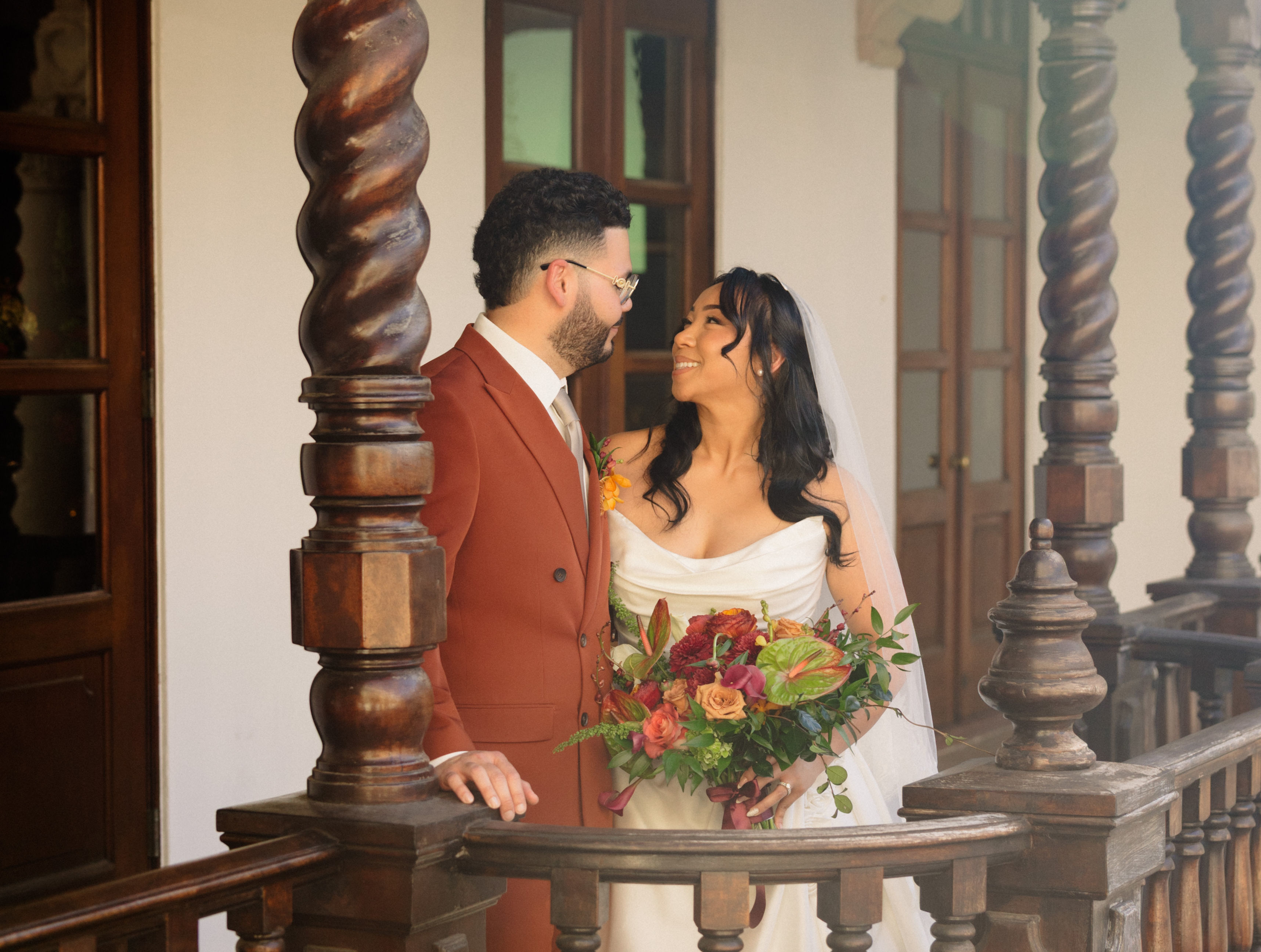 Bride and groom in fall wedding attire, with the groom wearing a terracotta suit and the bride holding a warm-toned autumn bouquet.