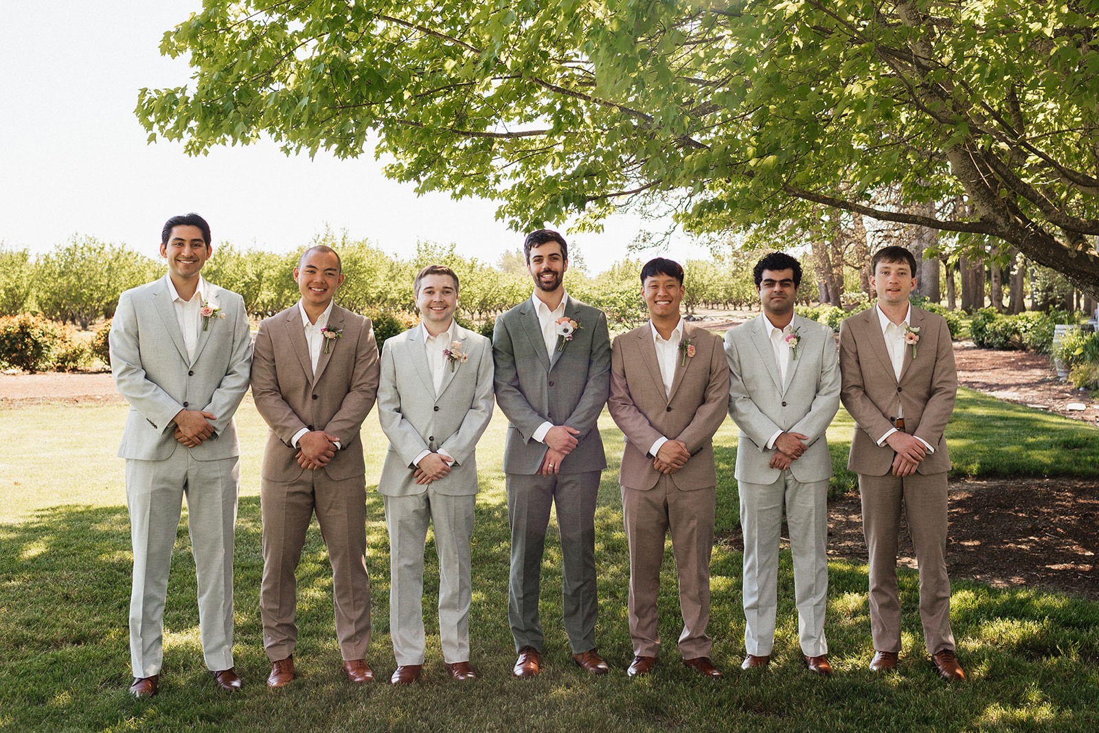 Seven groomsmen standing outdoors beneath a tree in mismatched light gray, taupe, and charcoal suits with white shirts and floral boutonnieres.