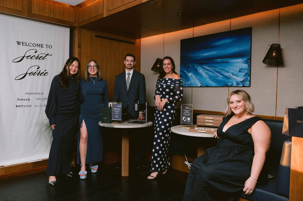 Five guests dressed in winter wedding attire pose at a Secret Soirée event beside a welcome banner and display tables.