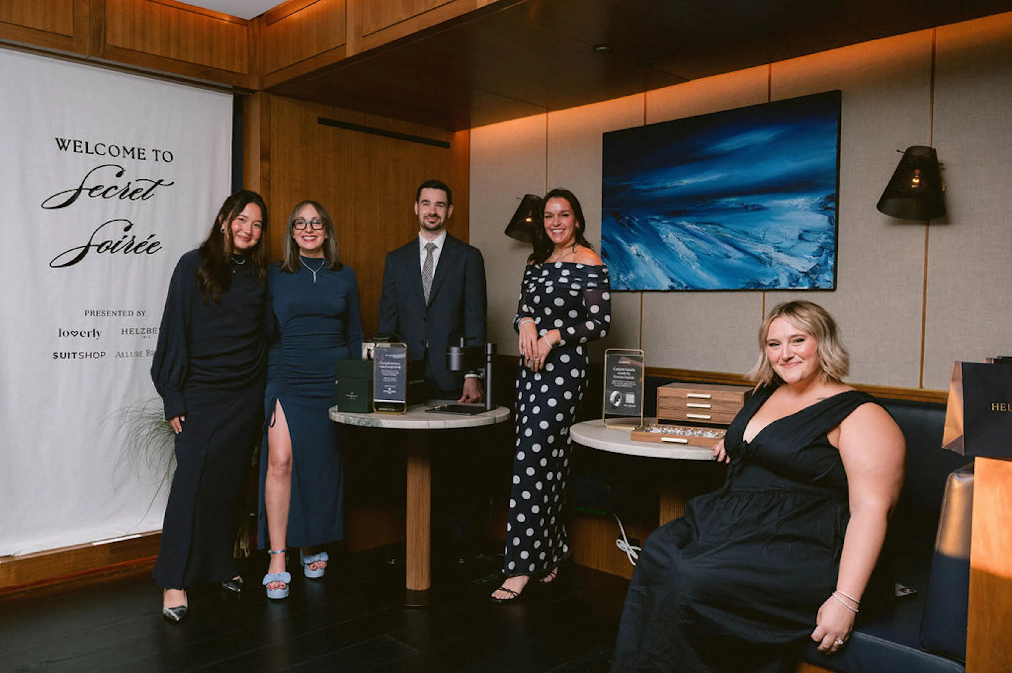 Five guests dressed in winter wedding attire pose at a Secret Soirée event beside a welcome banner and display tables.