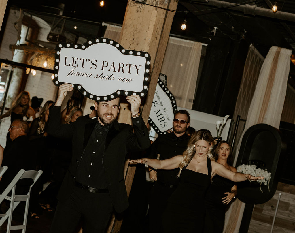Wedding guests dressed in black entering a reception, with one man holding a “Let’s Party” sign.