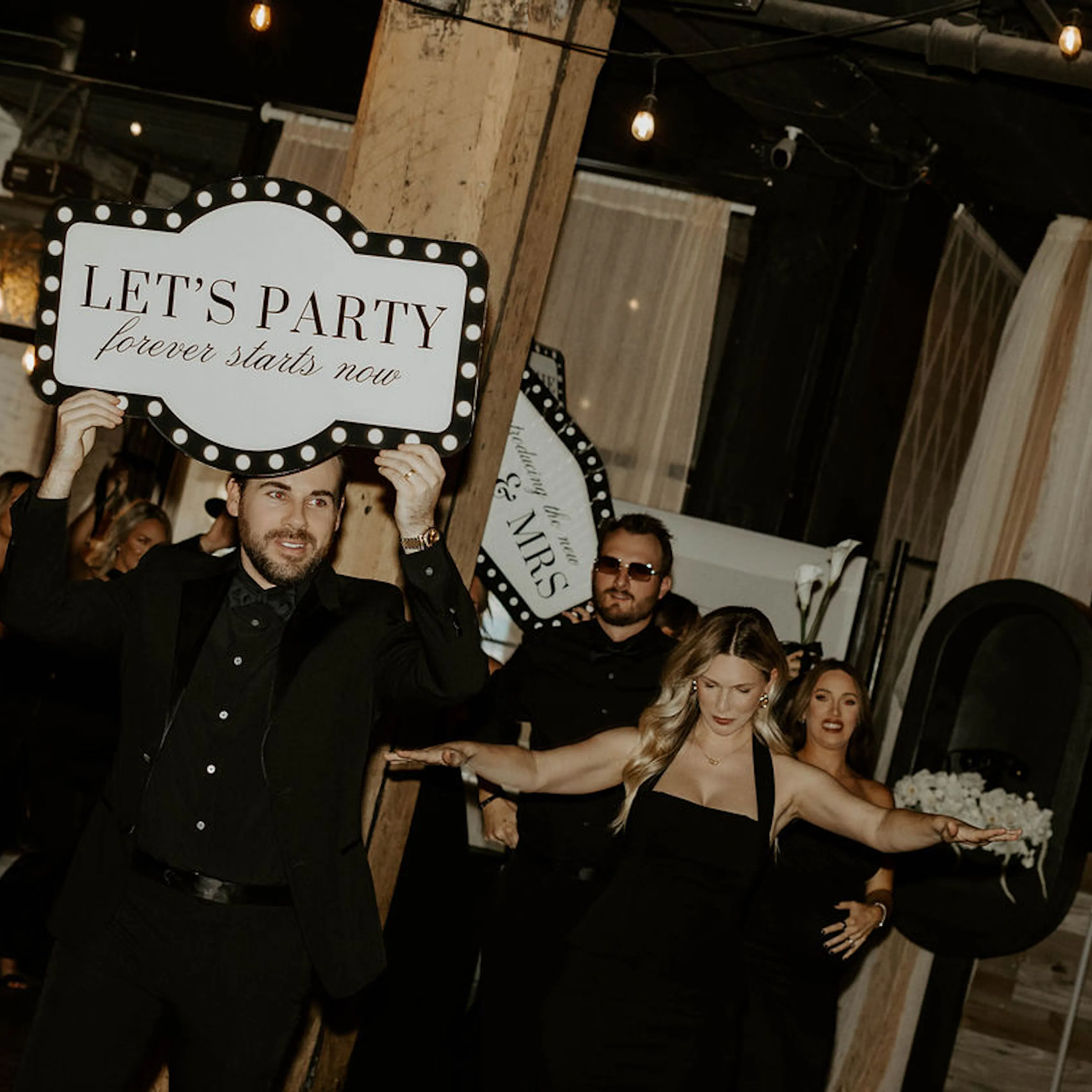 Wedding guests dressed in black entering a reception, with one man holding a “Let’s Party” sign.
