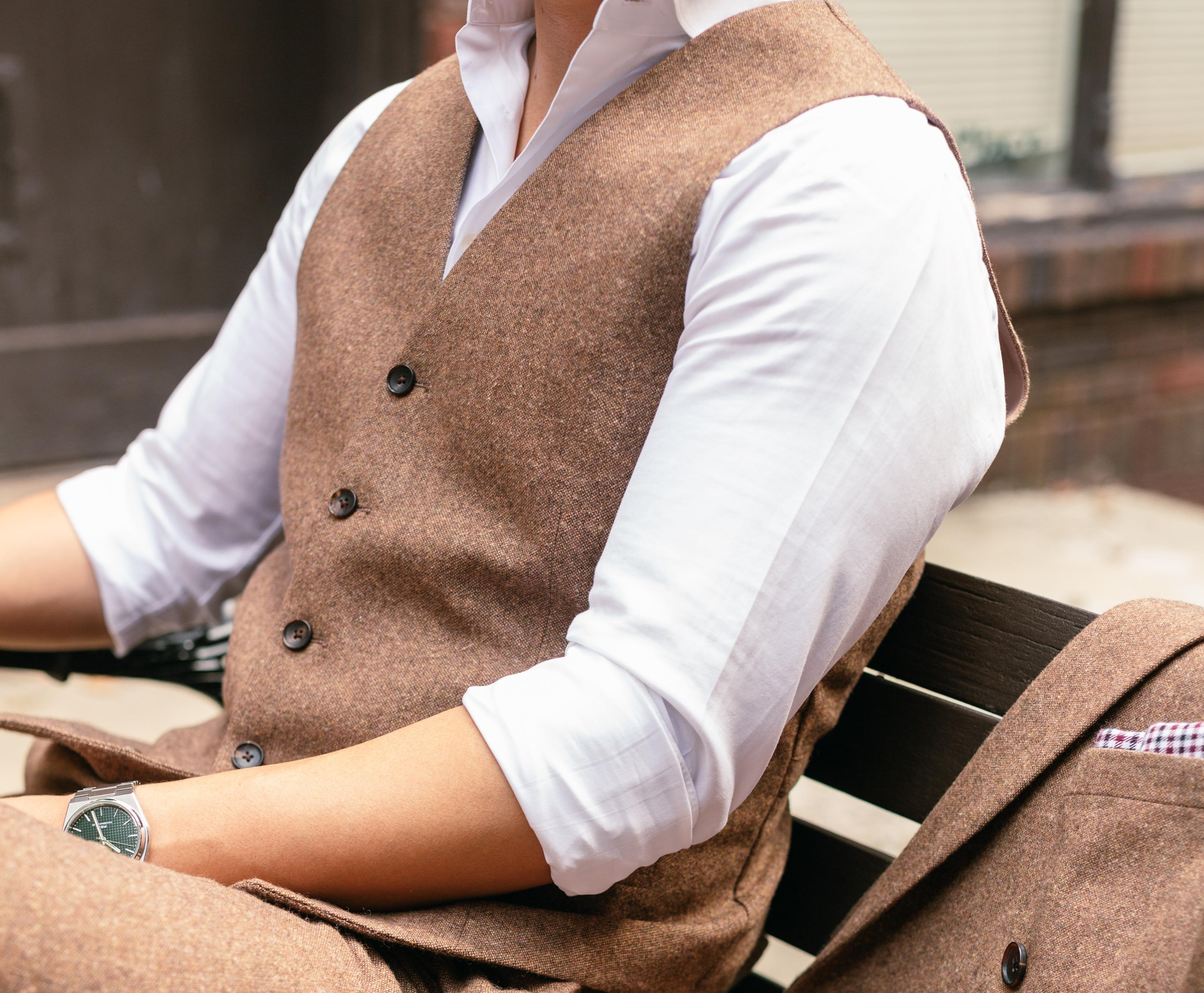Man wearing a brown waistcoat over a white dress shirt, seated with a matching brown suit jacket beside him.