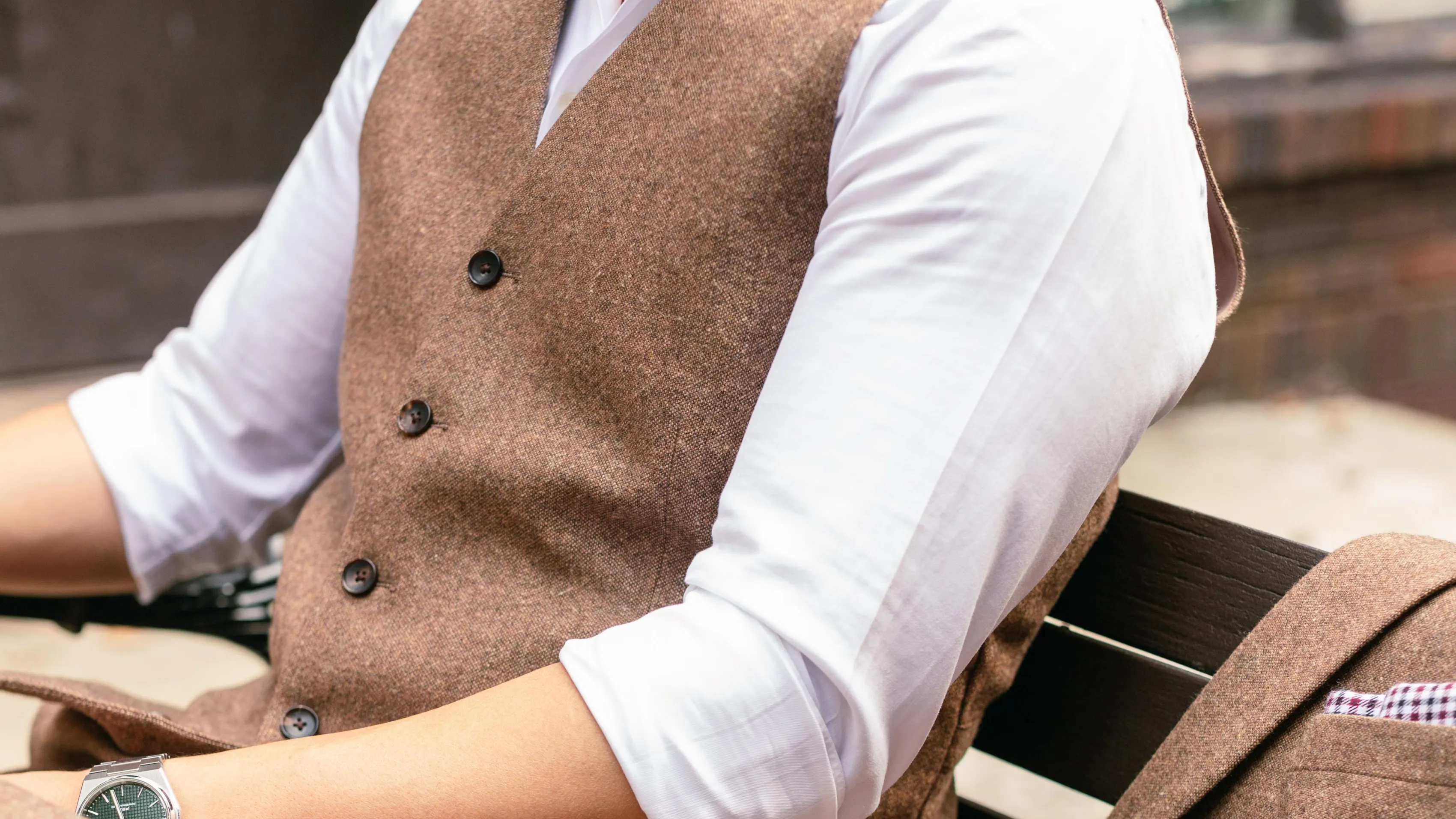 Man wearing a brown waistcoat over a white dress shirt, seated with a matching brown suit jacket beside him.