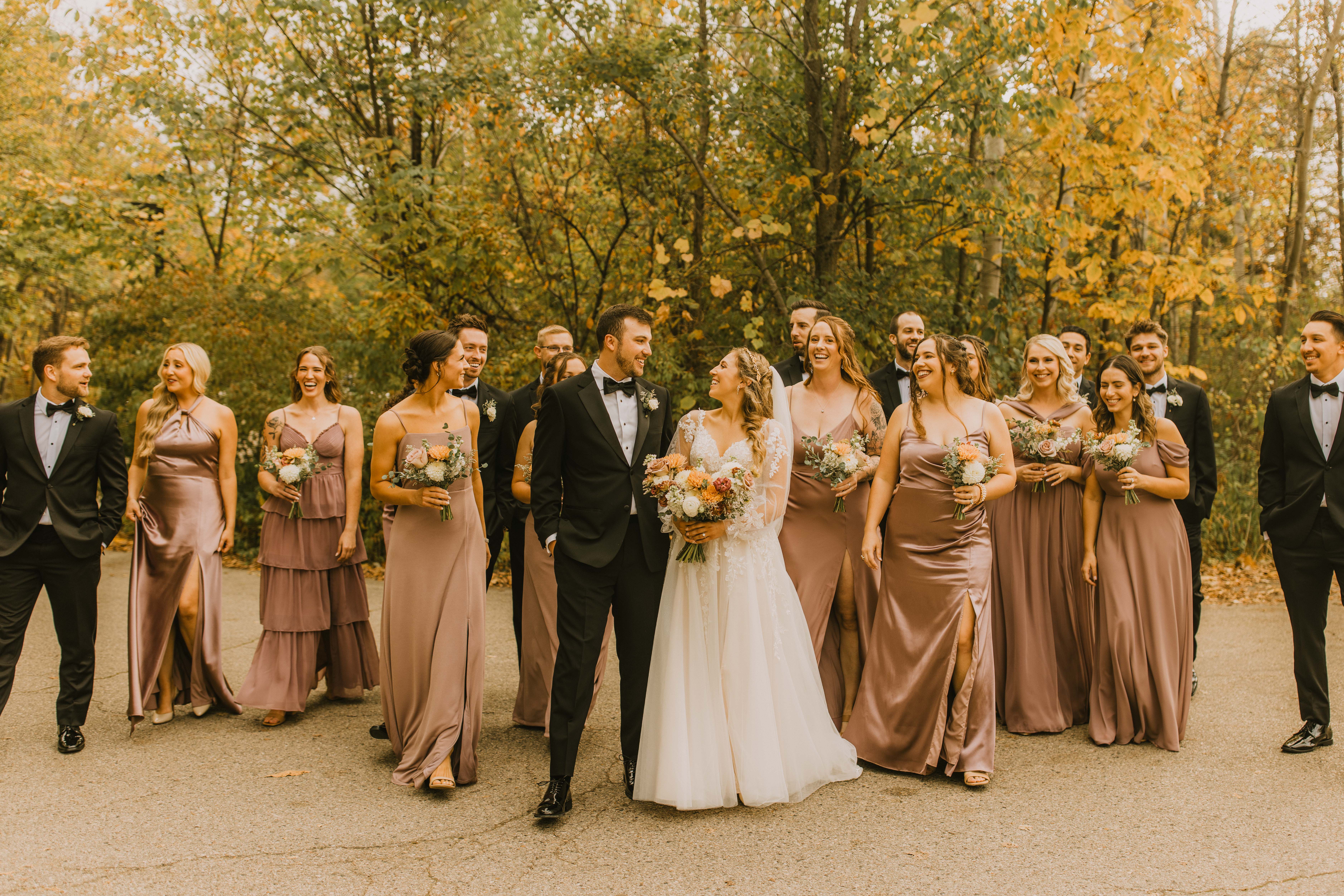 Bride and groom with their wedding party in black suits and mauve dresses surrounded by fall foliage.