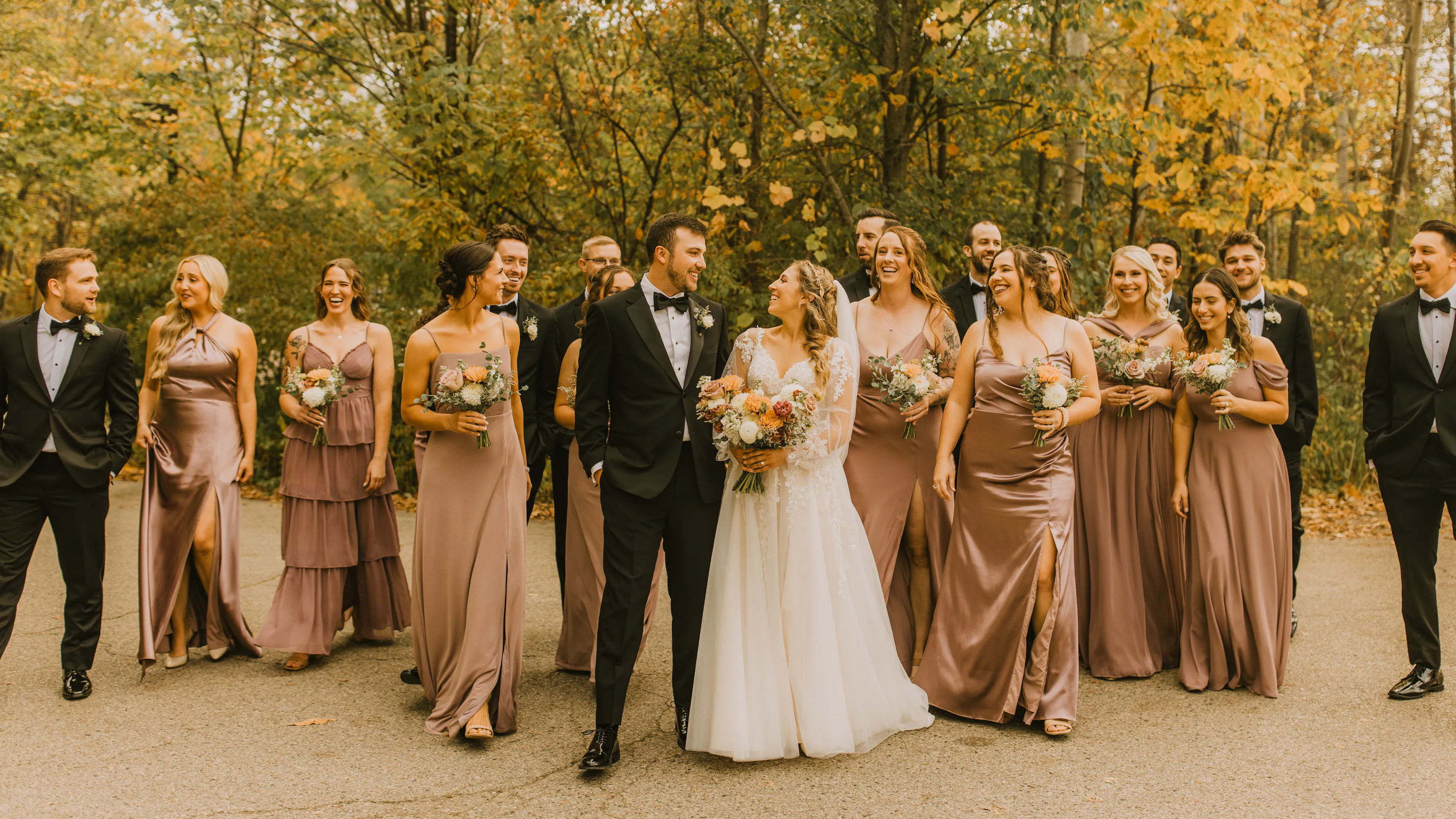 Bride and groom with their wedding party in black suits and mauve dresses surrounded by fall foliage.