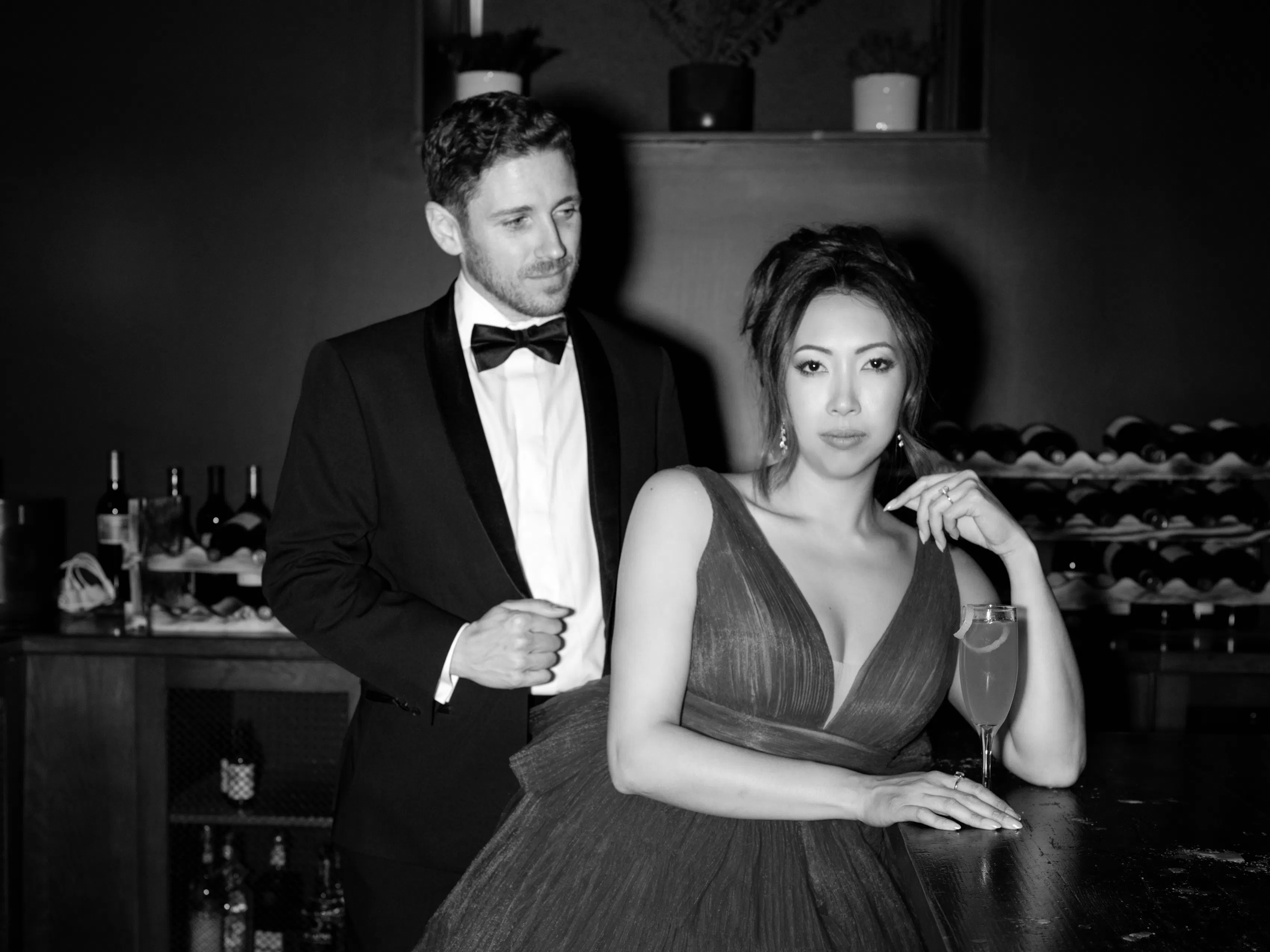 Black-and-white photo of a formally dressed couple, with a man in a tuxedo standing beside a woman in an evening gown holding a drink.