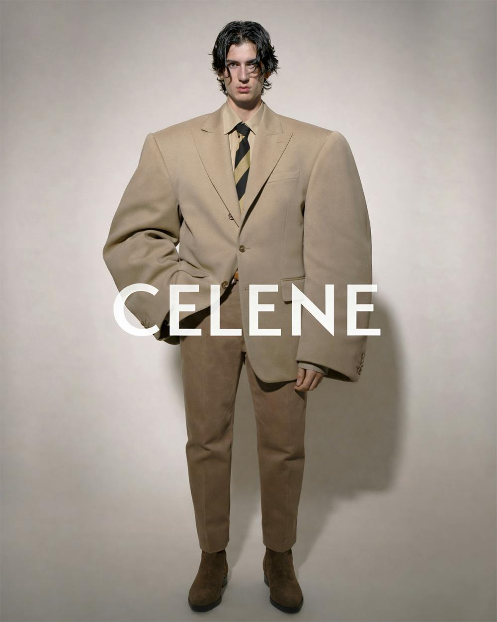 CELINE Model in tan suit with black-and-yellow striped tie and brown boots against beige background.