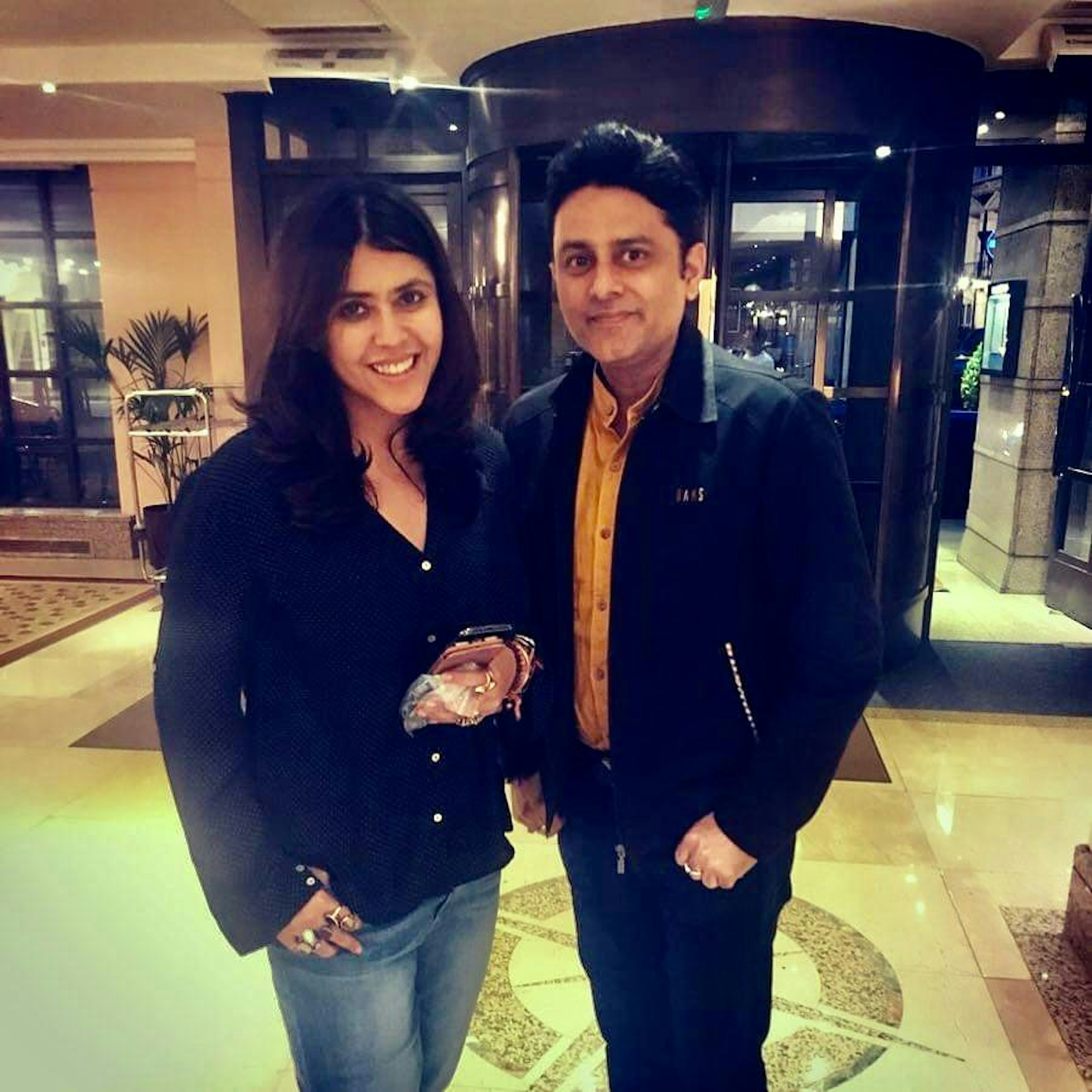 Ekta Kapoor wearing a dark blue shirt and jeans standing next to Dr. Sundeep Kochar in a black jacket and yellow shirt in a hotel lobby.