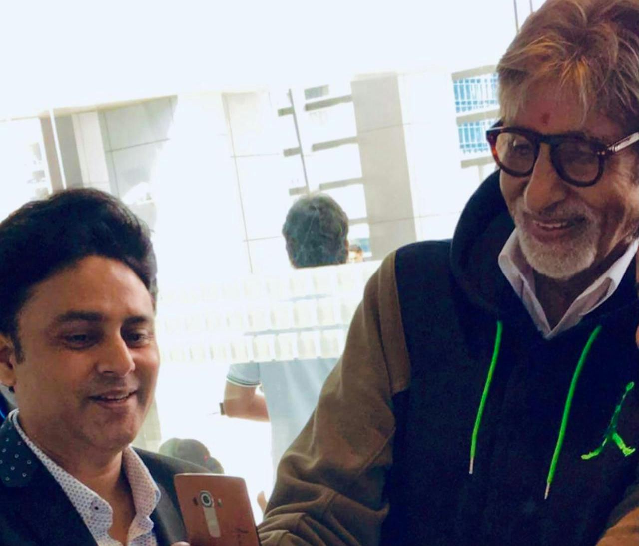 Amitabh Bachchan in a black Jordan hoodie and glasses looking at a phone alongside Dr. Sundeep Kochar.