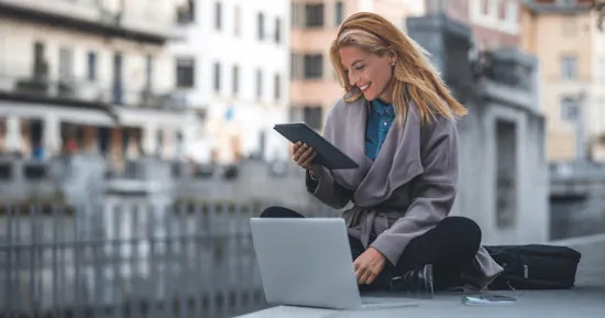 Woman reading on her tablet sitting in front of a laptop.
