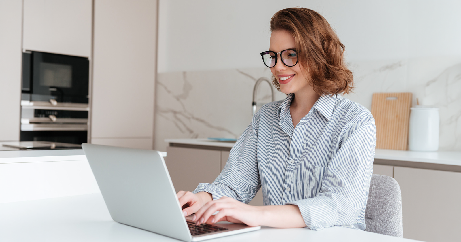 Smiling woman with glasses working on a laptop in a modern kitchen.