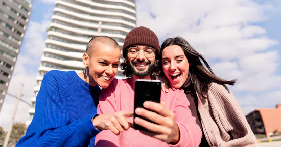 Three smiling people standing outdoors in front of modern high-rise buildings while looking at a smartphone together.