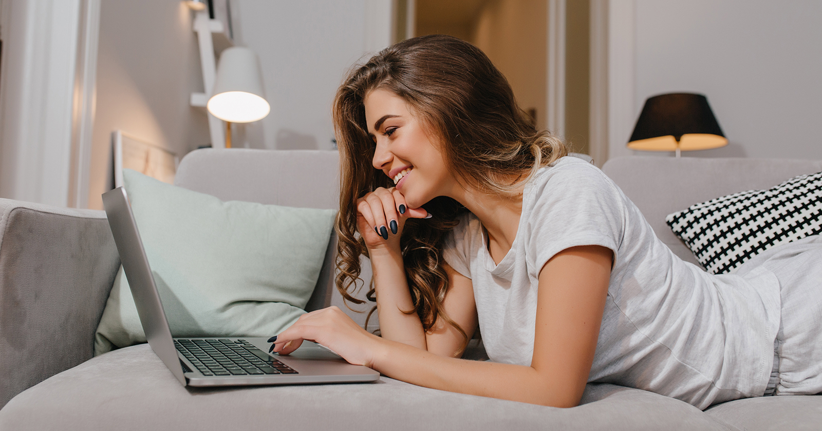 Young woman lying on her sofa at home, smiling while browsing on her laptop.