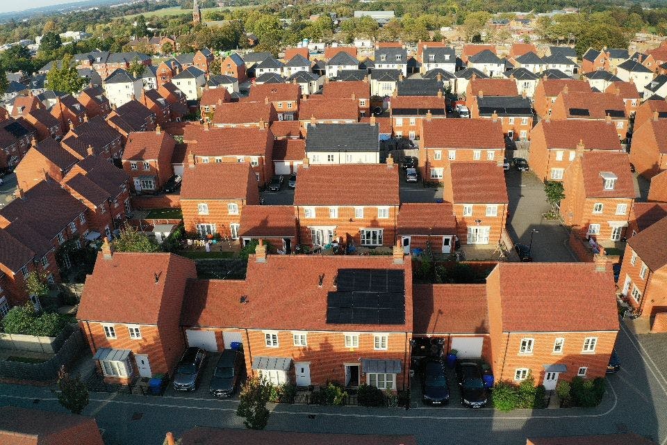 A bird's-eye view of dozens of red-brick and white houses, some of which have black solar panels. A park is in the background