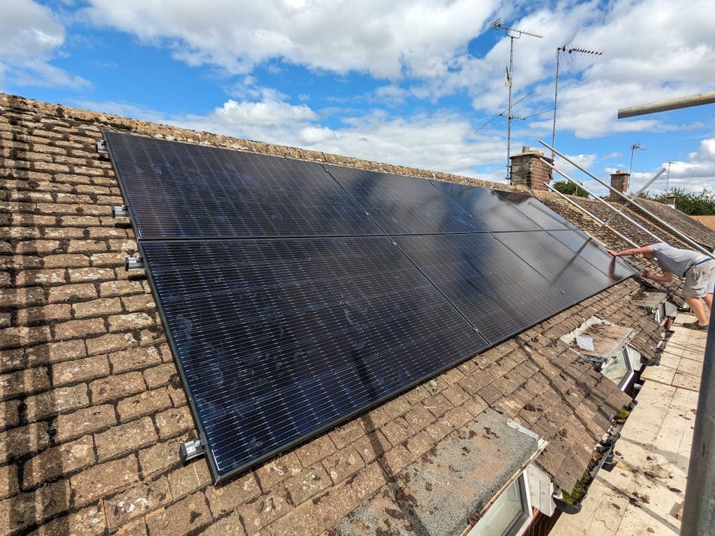 a photo of black solar panels being installed on a tiled, light brown roof under a blue sky with clouds. an installer is leaning on one of the solar panels