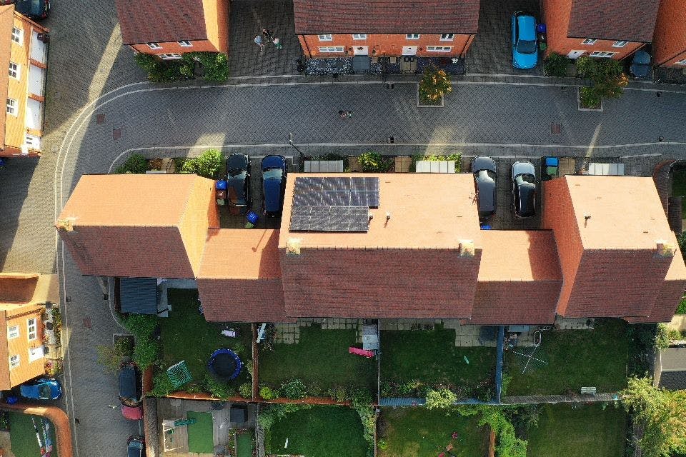 A bird's-eye view of black solar panels on a terracotta roof of a detached house, near other houses and trees, with gardens and a street also visible