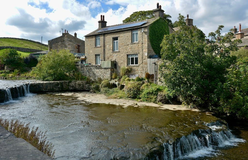 A stream flowing past a house in the UK countryside, solar panels on the house's roof, green hills in the background