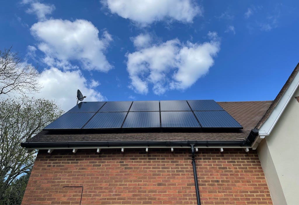 Black solar panels on a brown roof of a brick house. A satellite dish is also on the roof, which sits under a blue sky dotted with white clouds, with a tree in the background