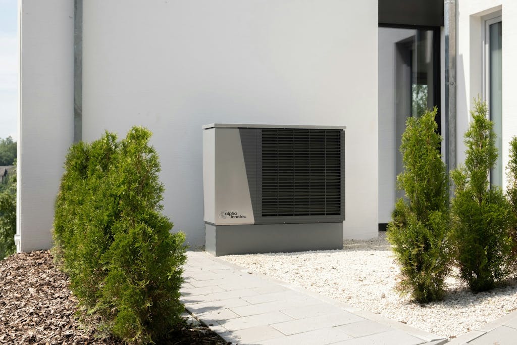 Grey and black heat pump against a blank residential wall, on a bed of white stones and shrubs