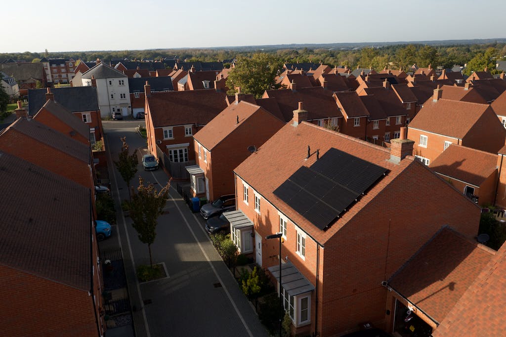 Ten black monocrystalline solar panels on a red rooftop in a UK housing estate, lots of red houses and trees in the background