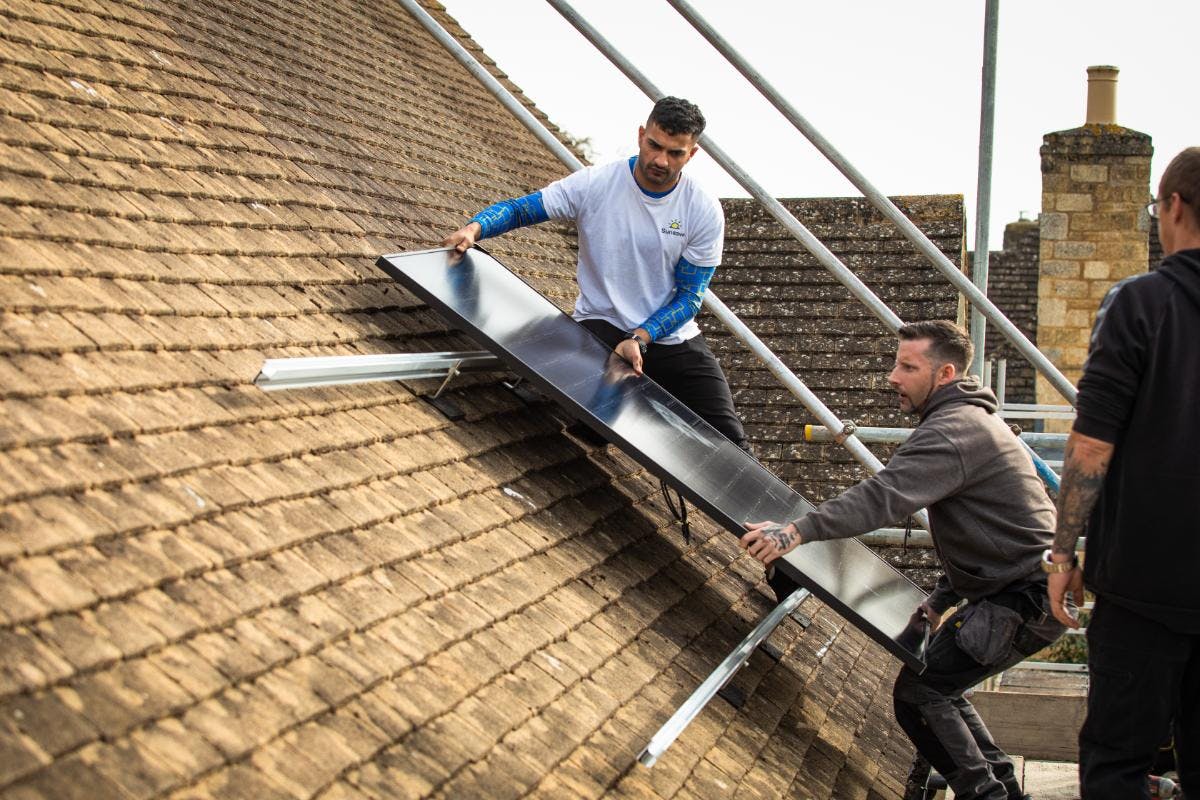 Two tradespeople on a roof affixing a black solar panel, with a third tradesperson on the right of the image