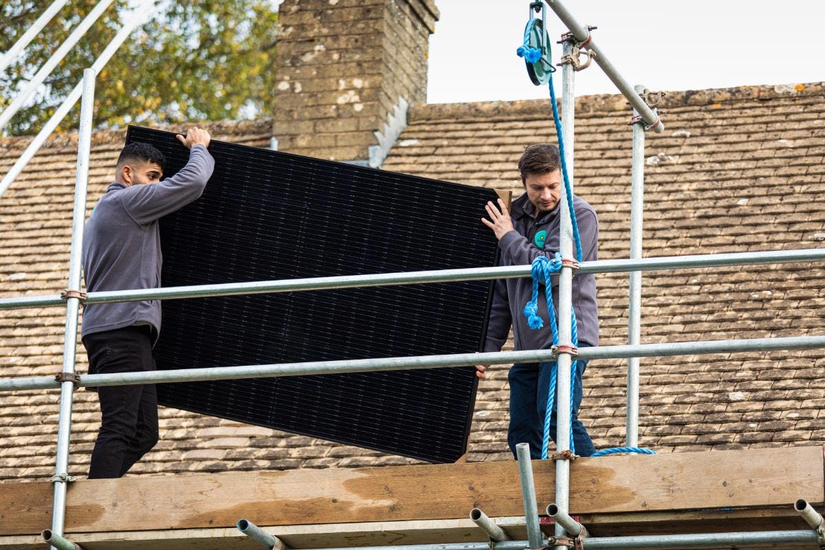 Two installers carrying a black solar panel along some scaffolding, in front of a house's roof