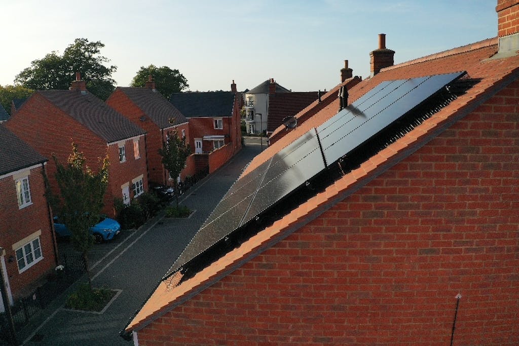 Black solar panels on a terracotta roof, with other houses and trees on the left, under a blue sky