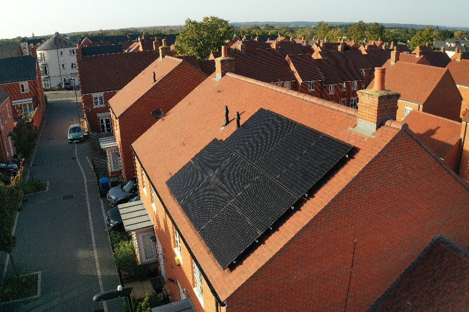 Black solar panels on a terracotta roof of a detached house, in a neighbourhood near other houses and trees, under a pale blue sky