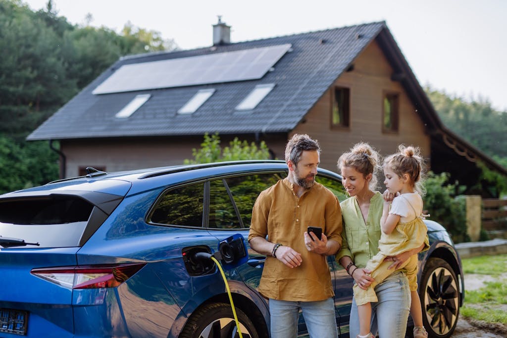 Man showing his phone to woman holding a child. They're all in front of an electric car, which is charging, and a house with solar panels on the grey roof