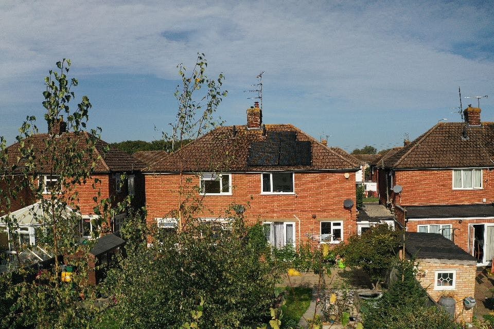 Three red-brick houses, face on, with trees in the foreground. The middle house has black solar panels on its brown roof, under a lightly cloudy blue sky