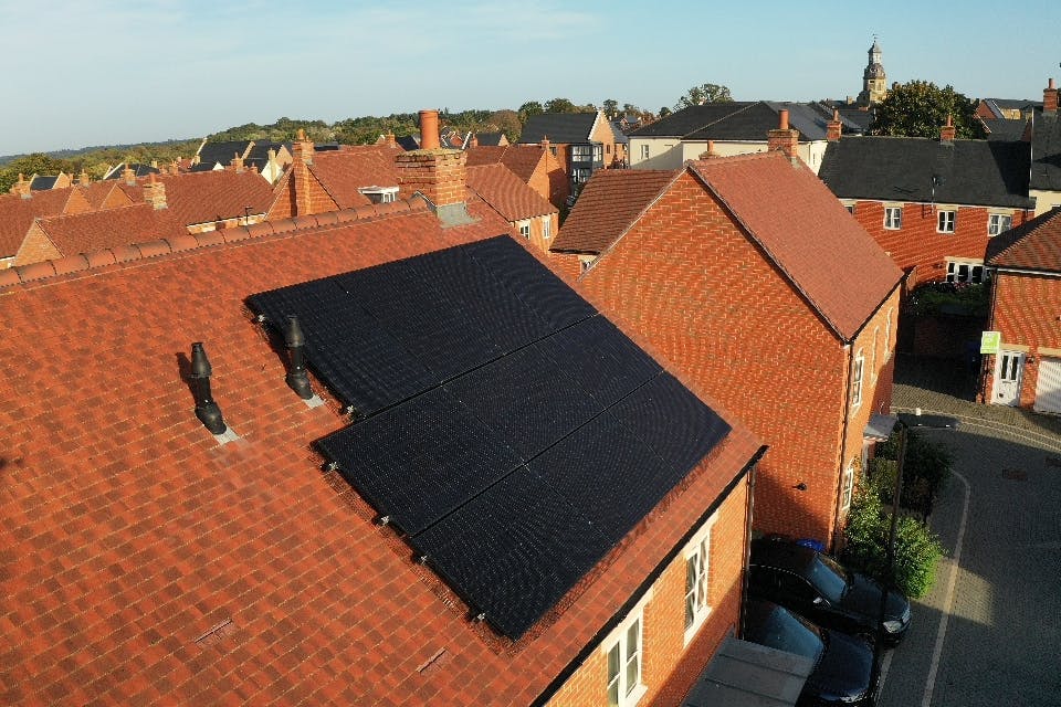 Black solar panels on a terracotta roof of a detached house, in a neighbourhood near other houses and trees, under a pale blue sky