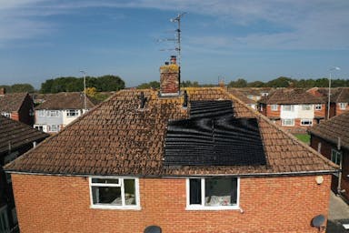 A drone's view of black solar panels on a terracotta roof of a detached house, near other houses, under a blue sky with wispy white clouds