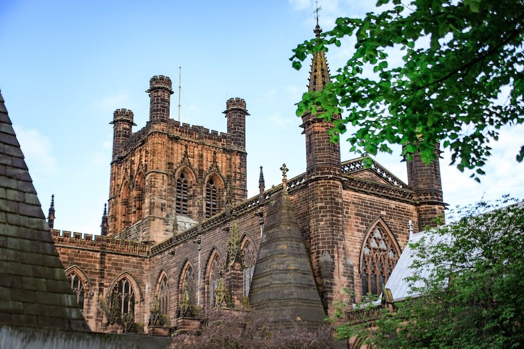 Chester Cathedral in the daytime from ground level, foliage in the top right corner, blue sky in background