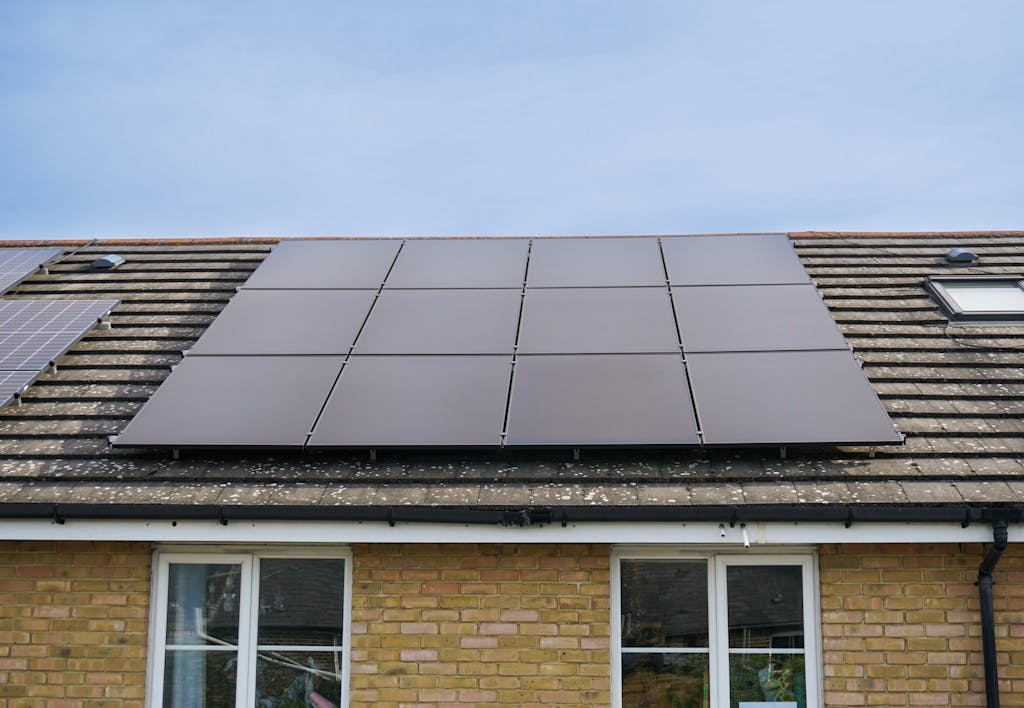 black solar panels on a brown roof, next to blue solar panels on a different home's roof, under a blue sky