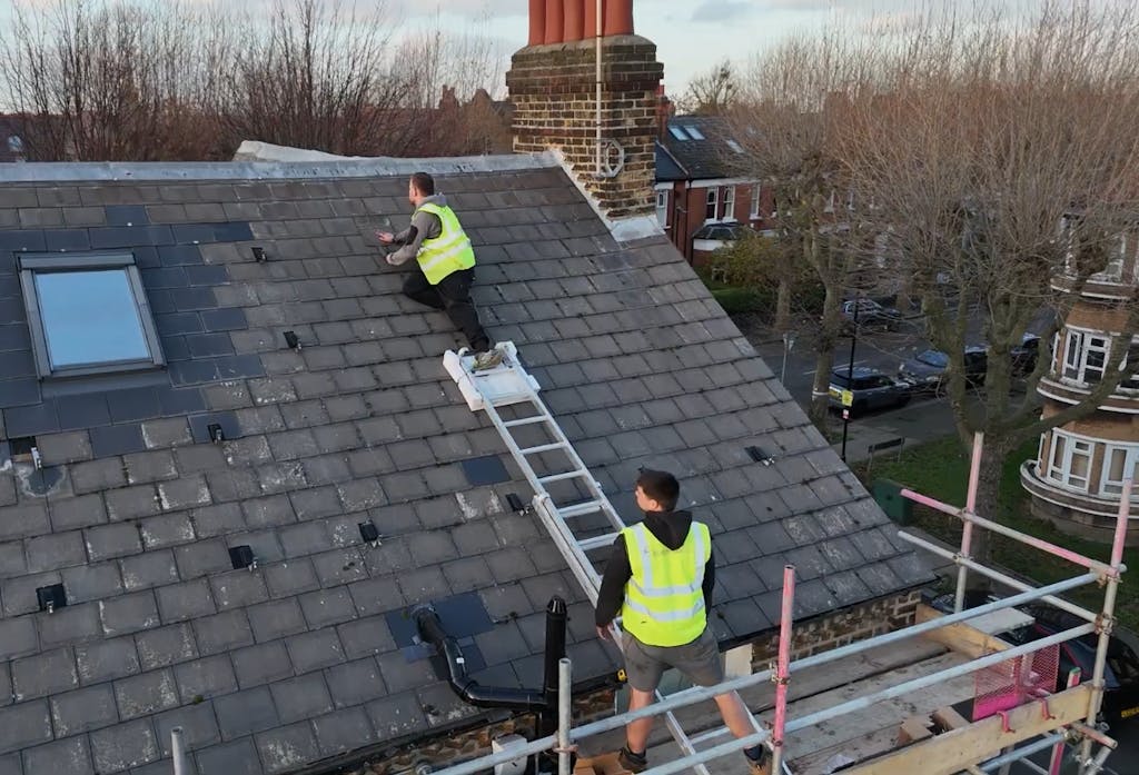 two installers in high-vis vests on a brown roof, climbing a ladder in the middle of a solar panel installation