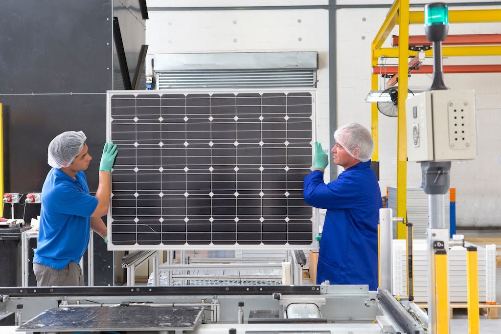 Two factory workers in blue shirts holding a black monocrystalline solar panel