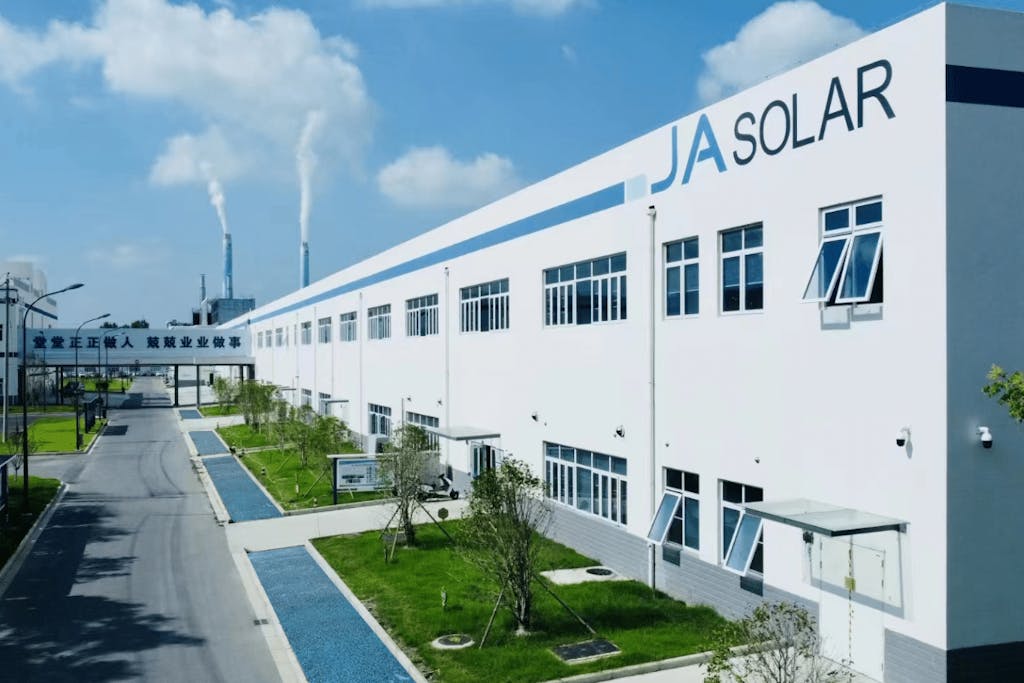 A white factory with JA Solar written on the outside, against a blue sky