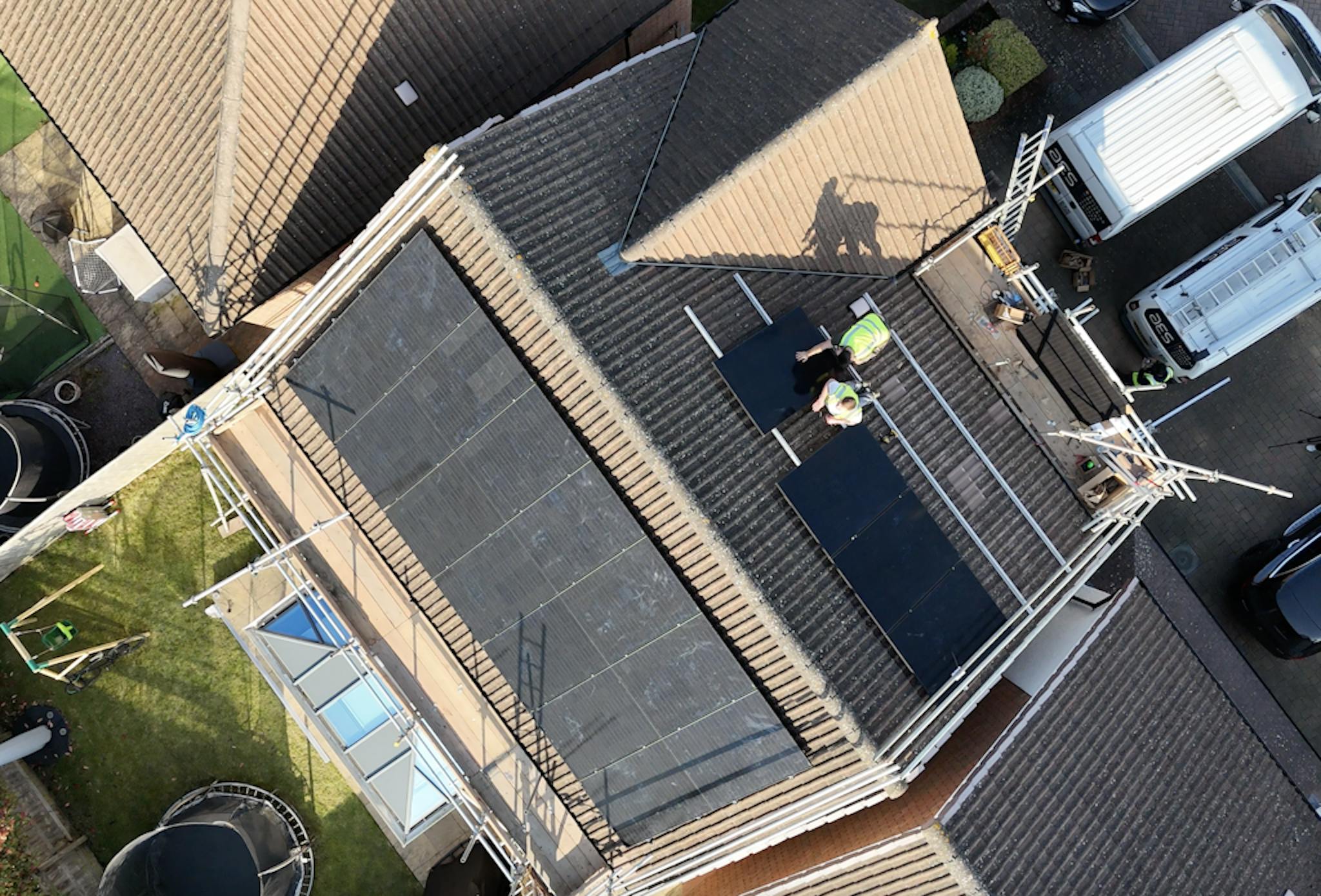 a brown roof with black solar panels, and two installers in hi-vis jackets installing more panels