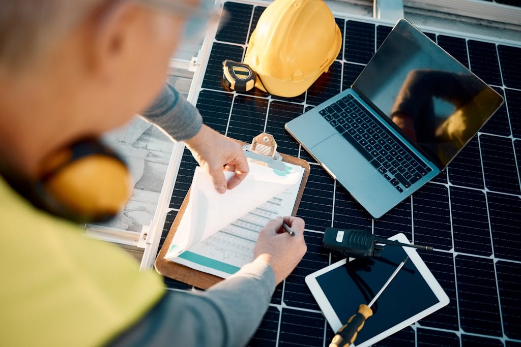 A person making notes on a clipboard while resting on a solar panel. A laptop, tools, hardhat, walkie talkie, and tablet are also on the panel